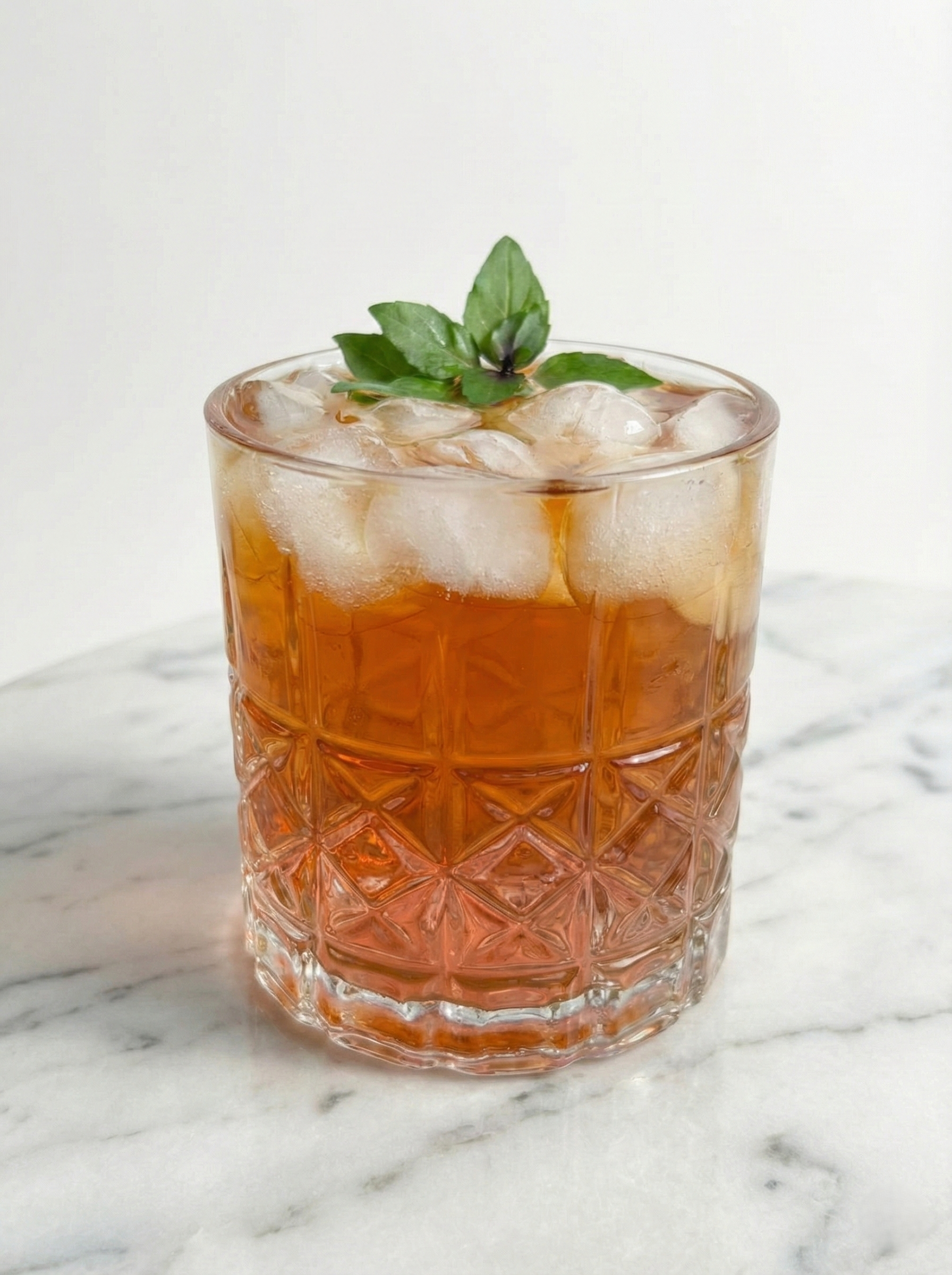 A photograph of a cocktail in a crystal-cut rocks glass filled with crushed ice and an amber liquid, garnished with a sprig of fresh basil. The glass sits on a white marble surface against a plain white background