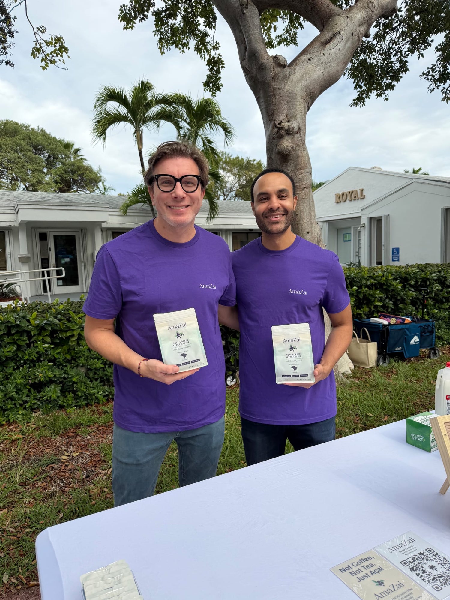 The two co-founders of Amazai smile proudly, holding bags of their product in front of the white Minnie Childhood Curated venue in Miami Beach.