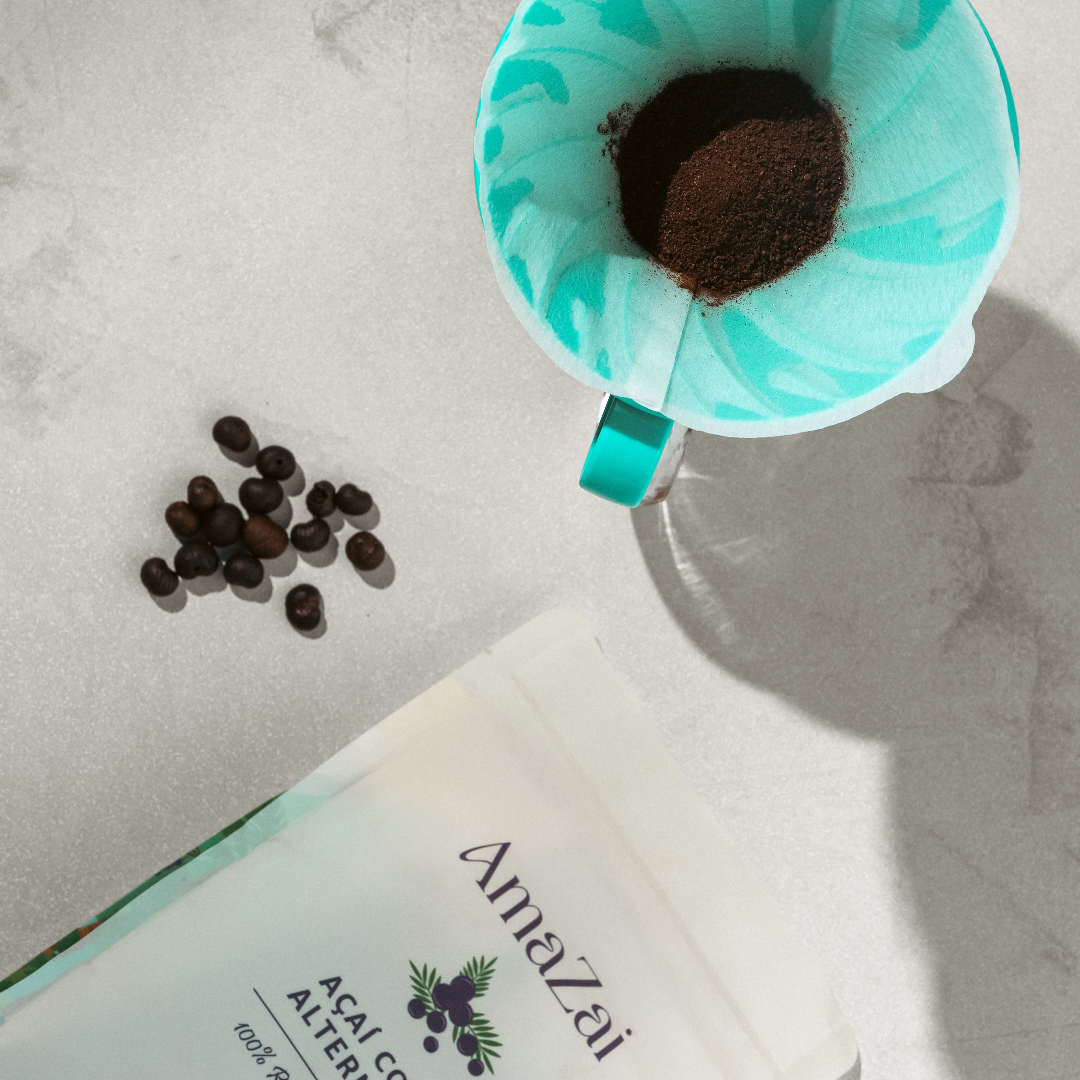 Turquoise coffee filter with coffee beans on a white surface next to an Amazai coffee alternative package.